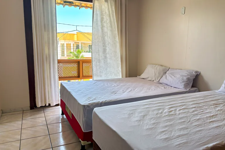 Quarto com duas camas e porta janela aberta para varanda com ceu azul