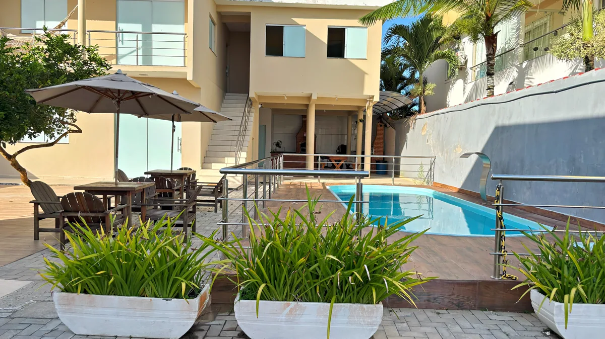 Area de lazer do residencial de dia, com piscina compartilhada, cadeiras de descanso, guarda-sol e o sobrado ao fundo